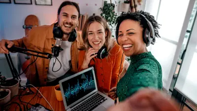 Foto von drei jungen Leuten in einem improvisierten Podcast-Studio, die in die Kamera lächeln