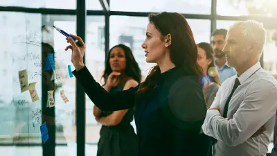 Image of a group of business people standing in front of a whiteboard with a woman in front, pointing at the board with a whiteboard marker
